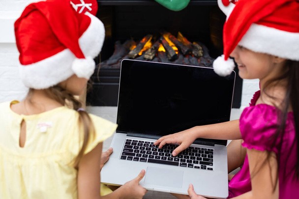 δύο μικρά κορίτσια, αδελφές, φίλες, παίζουν παιχνίδια, βλέπουν κινούμενα σχέδια σε ένα φορητό υπολογιστή το βράδυ των Χριστουγέννων κοντά στο τζάκι - Φωτογραφία, εικόνα