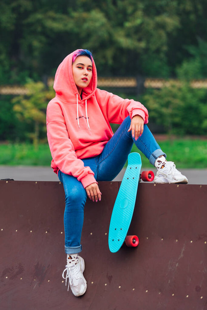 Portrait of a trendy pretty young girl with short colored hair and nose piercing dressed in pink hoodie and jeans sitting next to the skateboard court with her blue plastic skateboard. - Photo, Image