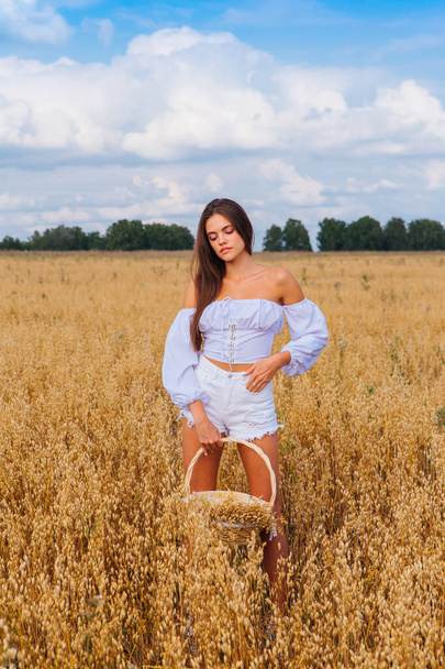 Rural Countryside Scene. Young beautiful woman with long hair dressed in white at golden oat field holding basket with ears of oats. - Photo, image