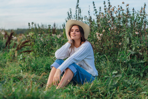 Jonge mooie brunette vrouw gekleed in een witte trui, jeans en cowboy stro hoed zittend op groen gras, glimlachen en lachen tijdens zonsondergang - Foto, afbeelding