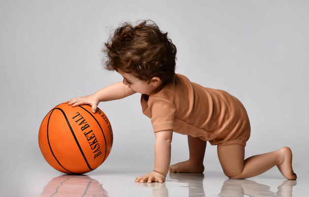 Baby boy dressed in a bodysuit with interest plays a basketball on a gray background. - Fotó, kép