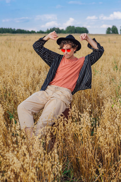 Rural Countryside Scene. Tall handsome man dressed in a black shirt, black hat and pink sunglasses sitting on a brown vintage leather suitcase at golden oat field. Summer landscape with blue sky - Photo, image