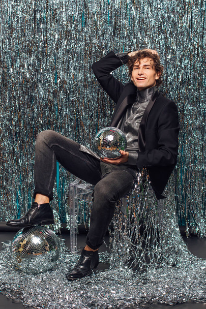 Tall attractive man sitting on the chair over the sparkle background holding mirror disco ball. Party concept. - 写真・画像