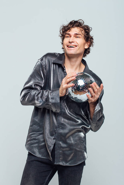 Portrait of tall attractive man with a mirror disco ball on a white background. Handsome man posing with a shiny disco ball in studio. - 写真・画像