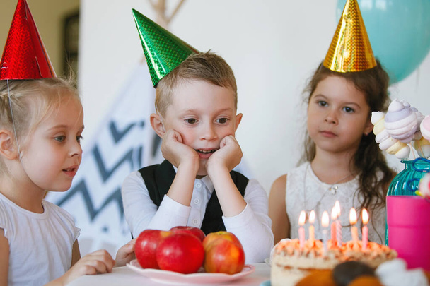 Children celebrate their birthday with a cake with candles. Multi-colored caps, red apples, bright eyes of boys and girls. Happy childhood. Sweet happiness. Selective focus. - Photo, Image
