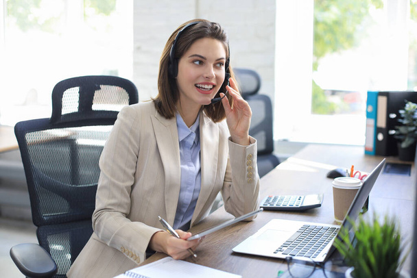 Cheerful female manager sitting at office desk and performing corporate tasks using wireless connection on digital gadgets - 写真・画像