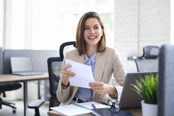 Attractive cheerful business woman checking paper documents in office, working on laptop - Foto, Imagem