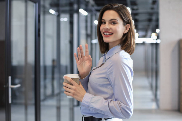 Businesswoman walking along the office corridor with paper cup and waving hand by greeting you - 写真・画像