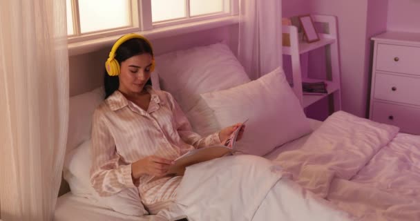 Kaunis nuori nainen kuuntelee musiikkia lukiessaan lehteä sängyssä - Materiaali, video