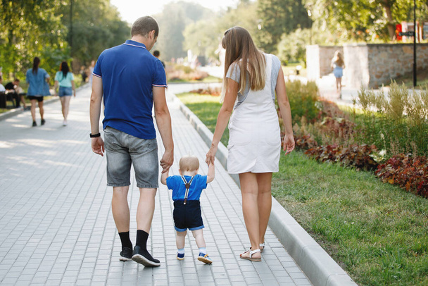 Le père, la mère et le fils de la famille marchent sur le trottoir dans le parc de la ville en été. T-shirts bleus, journée ensoleillée, idylle familiale. Bonne enfance. Concentration sélective. Vue arrière. - Photo, image