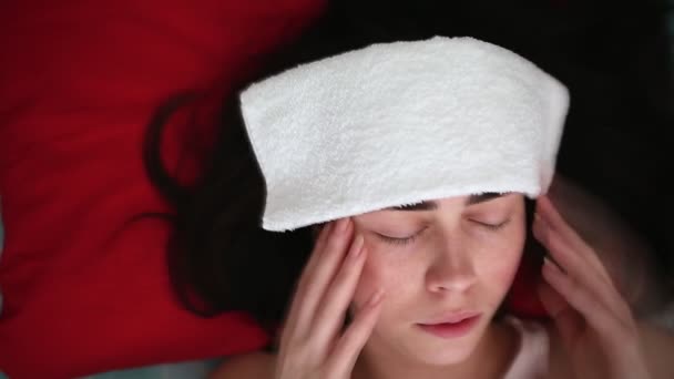 Eine junge Frau liegt mit einem Tuch auf der Stirn im Bett und hat starke Schmerzen im Kopf. Porträt aus nächster Nähe. Der Blick von oben. Das Konzept der Laptopschmerzen und Erkrankungen des Kopfes - Filmmaterial, Video
