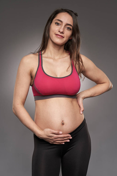 Активная беременная женщина в спортивных брюках и лифчике на сером фоне - Фото, изображение