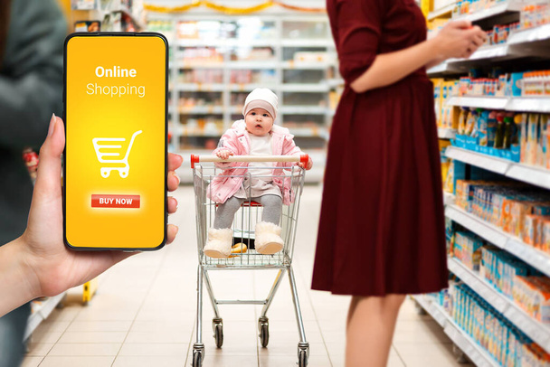 Nakupovat. Roztomilé dítě sedící v nákupním vozíku v supermarketu, vedle jeho mámy. Ruka na levé straně drží smartphone. Koncept online nakupování. - Fotografie, Obrázek