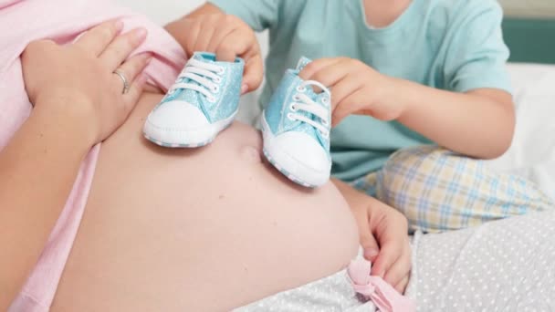 妊娠中の母親の大きな腹で赤ちゃんの靴で遊んでいる男の子の閉鎖。明るい子供と赤ちゃんを期待する家族の幸せの概念 - 映像、動画