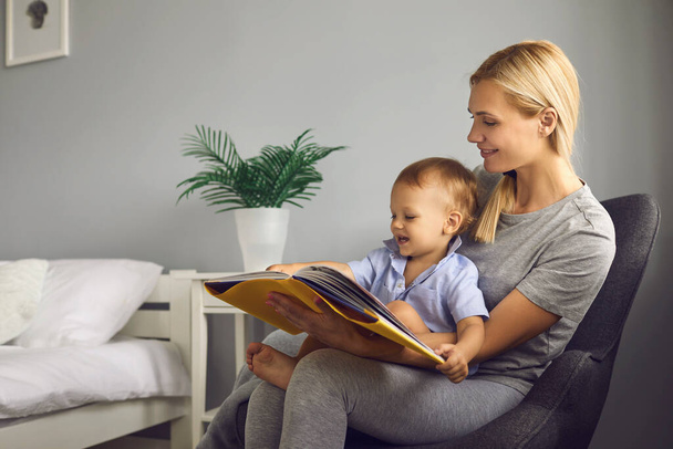 Mother reads a childrens book to her little toddler son sitting in a chair in a cozy bedroom. - Fotografie, Obrázek