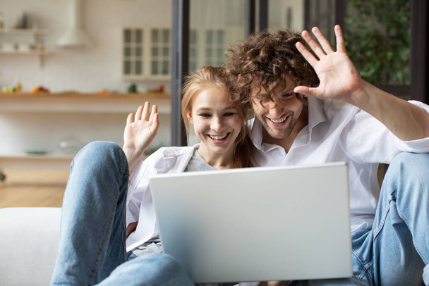 Happy young couple relaxing and smiling on a couch at home and making a video call to friends on laptop. Remote communication. - Φωτογραφία, εικόνα