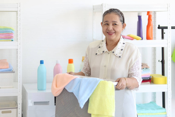 Glücklich lächelnde asiatische Seniorin mit Wäschekorb in der Waschküche mit Waschmaschine, Oma mit Hausarbeit und Wäschewaschen im Haus. - Foto, Bild