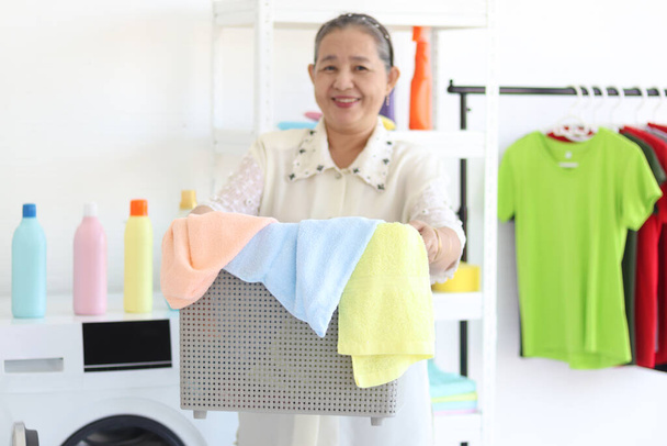 洗濯機付きの洗濯室で洗濯をするための衣類バスケットを持っている幸せな笑顔アジアの高齢女性の主婦,家事や家の中で衣類を掃除するおばあちゃん. - 写真・画像