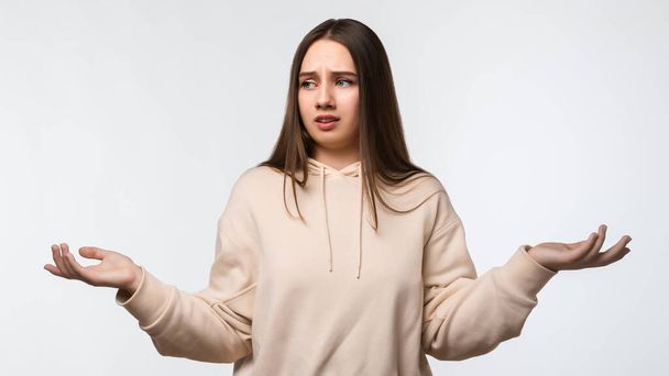 Studio shot of Insecure puzzled young female with long chestnut hair, spreads hands with doubt, wearing stylish beige hoodie. Concept of doubt, uncertainty and confusion - 写真・画像