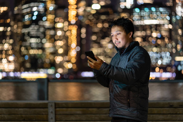 Asiatischer Mann im Mantel-Anzug mit Smartphone und Lächeln-Aktion über dem Foto verschwommenen Bokeh von New York Stadtbild neben dem East River, USA Innenstadt, Karriere überall Konzept - Foto, Bild