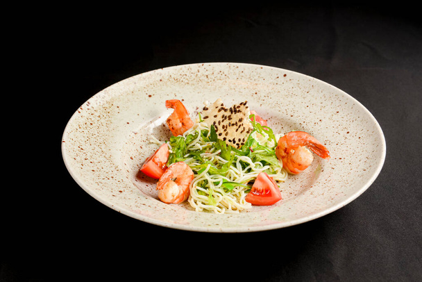 Rice noodles with tomatoes, fresh lettuce saladn and sesame seeds. Asian noodles salad on a plate over black background. Asian cuisine concept. - Foto, imagen