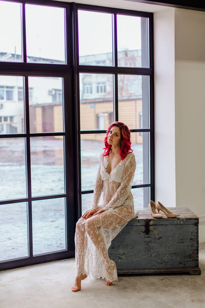 Attractive woman with pink hair in white light dress sitting on the old wooden box near large window. Lady in gentle lace peignoir sitting with bare feet in a room with concrete floor - Foto, imagen