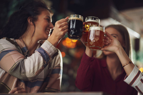 Three cheerful girlfriends are having fun time in pub. They are toasting. Focus on beer glasses. - Foto, Imagem
