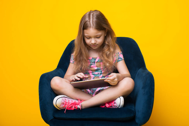 Adorable chica está usando una tableta mientras está sentado en un sillón en una pared de estudio amarillo - Foto, imagen