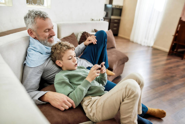 Children and modern technologies. Little boy grandson using smartphone while relaxing on sofa at home with his happy grandfather - Foto, afbeelding
