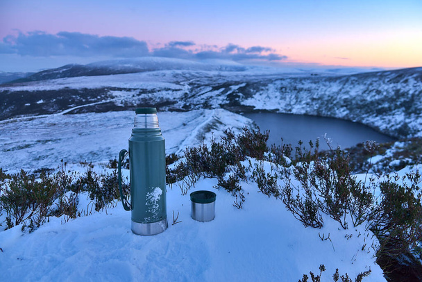 Thermos e una tazza di tè al vertice innevato e vista panoramica serale sul lago Lough Bray Upper visto da Eagles Crag, Ballylerane, Co. Wicklow, Irlanda. L'ora del tè durante le escursioni - Foto, immagini