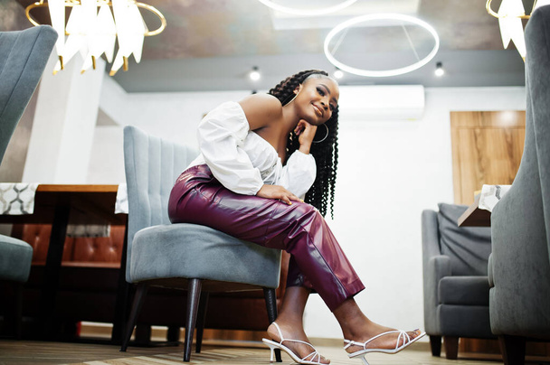 Beautiful african american woman in white blouse and red leather pants pose at restaurant. - 写真・画像