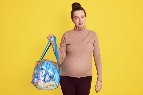 Pregnant woman posing against yellow wall with pout lips, being worry before going to maternity home, standing with bag with her stuff in hands, wearing casual sweater and leggins. - Φωτογραφία, εικόνα