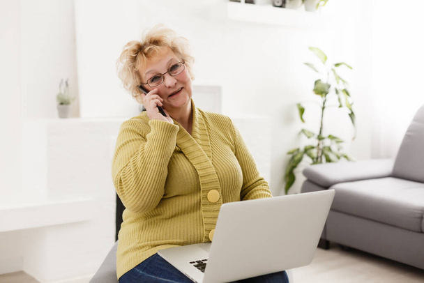 Attractive elderly woman sitting on couch at home, looking at camera, makes video call, communication distantly, modern wireless technologies concept - Φωτογραφία, εικόνα
