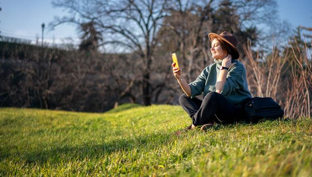 Schöne, fröhliche erwachsene Frau mit Filzhut und Pullover entspannt sich an einem Frühlingstag mit dem Smartphone in der Hand auf grünem Gras - Foto, Bild