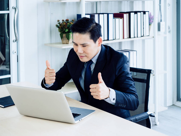 Jóképű ázsiai üzletember öltönyben nézi a képernyőt, és mutatja hüvelykujját a laptop számítógép, videohívás, találkozó online munka, videokonferencia. Társadalmi távolság vagy távközlés. - Fotó, kép