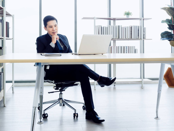 Knappe Aziatische zakenman in pak concentreren kijken op het scherm tijdens het werken met laptop computer op kantoor in de buurt van boekenplank en glazen venster. - Foto, afbeelding