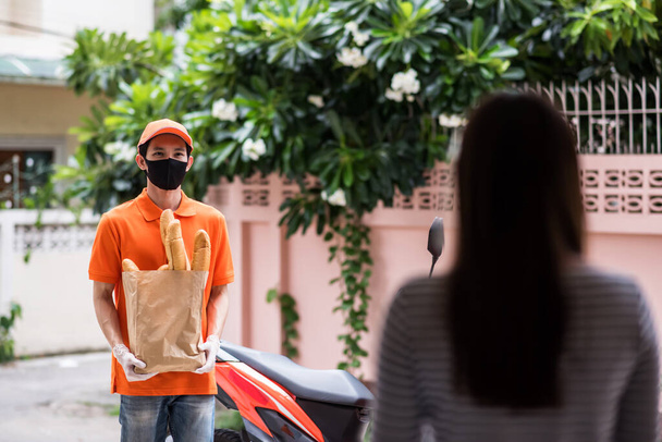 Un messager asiatique avec masque facial et gants en uniforme orange livre du pain à une femme devant chez elle. livraison express d'épicerie en moto pendant la période covid-19. Nouvelle activité normale. - Photo, image
