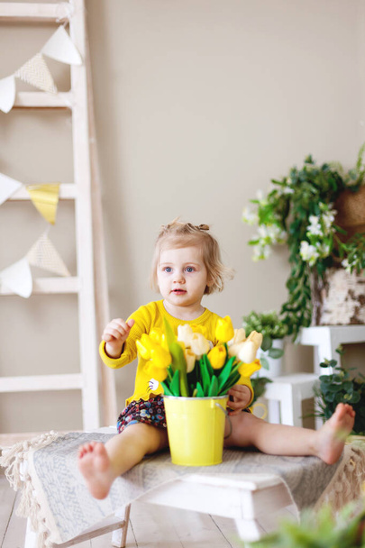 黄色のチューリップの束と黄色のドレスの小さな赤ちゃん。国際女性の日   - 写真・画像