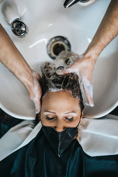 Hairdresser washing hair of a beautiful young adult woman in hair salon. She is wearing protective face mask as protection against virus pandemic. - Φωτογραφία, εικόνα