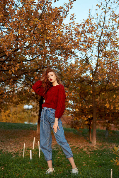 Redhead young woman in a red sweater walks in the park. Autumn beauty portrait of a fashionable Red-haired woman at sunset - Zdjęcie, obraz