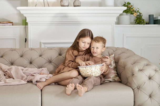 Brother and sister eating popcorn sitting at home on the couch. A boy and a girl relax have fun and watch a movie on TV - Фото, зображення