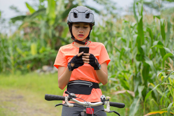 Ázsiai nő kerékpáros használni okostelefon, amikor lovaglás kerékpár a városi erdőben. - Fotó, kép
