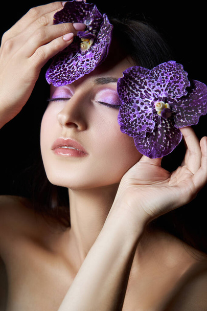 Beauty portrait of a nude woman with a purple flower in her hands on a dark background. Natural cosmetics, natural makeup - 写真・画像