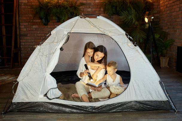 Mother reads a book of fairy tales for her children while sitting in a tent at night. Mom son and daughter reading a book with a flashlight in their hands - Foto, Imagem