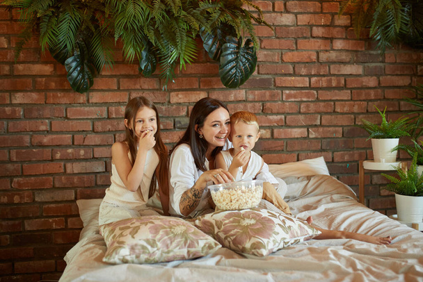Mom sits on the bed with her son and daughter and watch a movie. A woman, a boy and a girl eat popcorn while watching a movie in the bedroom. The family is resting at home on the weekend - Zdjęcie, obraz
