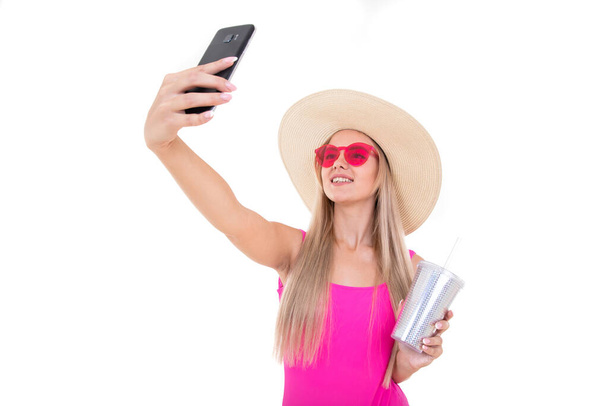 Portret młodej kobiety w różowym stroju kąpielowym i kapeluszu trzymającej szklankę i robiącej selfie - Zdjęcie, obraz