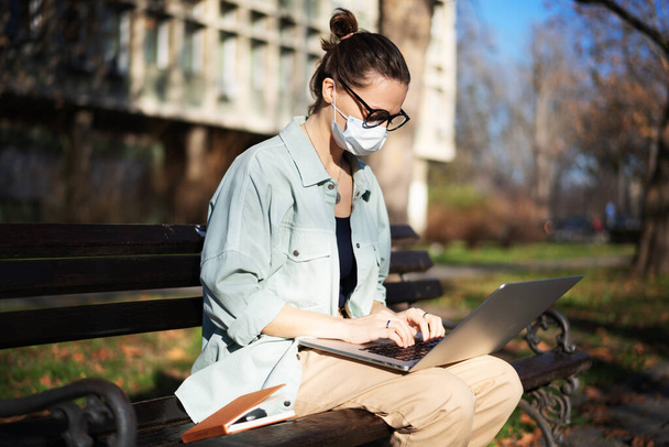 young woman student in a facial protective mask sitting on a bench in the park and typing on her laptop on a sunny day - Foto, afbeelding