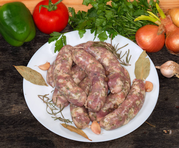 Сырые домашние свиные колбасы, приготовленные для дальнейшего приготовления на белом блюде на черной поверхности среди специй, зелени и овощей, полуфабрикатов - Фото, изображение