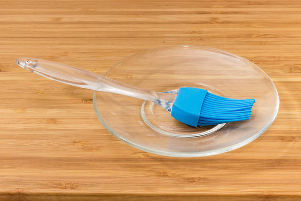 Gebäckpinsel mit blauen Silikonborsten und transparentem Kunststoffgriff auf einer Glasuntertasse auf einem hölzernen Bambusbrett - Foto, Bild