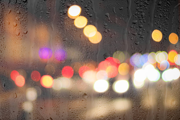 Luces nocturnas de la ciudad y faros de coche a través de vidrio lluvioso. Lluvia y mal tiempo húmedo concepto. - Foto, Imagen